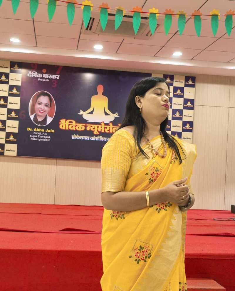 Dr. Abha Jain in a yellow saree on stage in front of the Dainik Bhaskar Vedic Numerology course banner